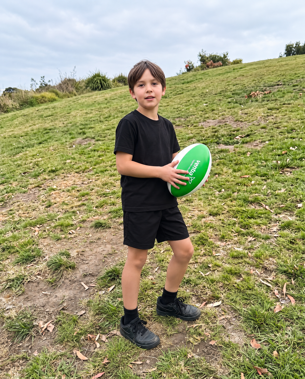 A young boy wearing a black t-shirt and black shorts stands on a grassy hill holding a green rugby ball with white text.