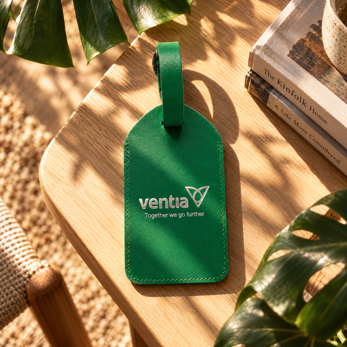 A green leather luggage tag with the brand name 'ventia' and the tagline 'Together we go further' printed in white, resting on a wooden table with natural leaf shadows and nearby books and plants.